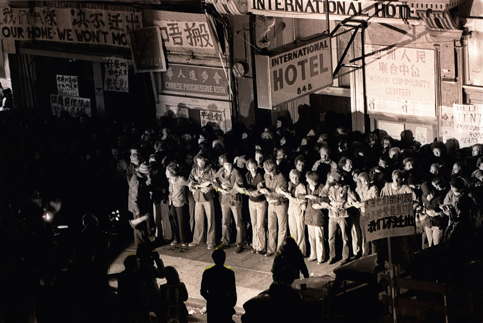 Black and white photo of hundreds of protesters linking arms in front of the I-Hotel in August of 1977 to protect tenants from eviction by police. Credit: Nancy Wong